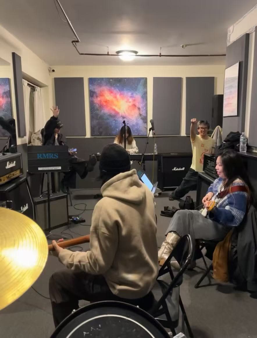 Band rehearsal in a studio with galaxy artwork on the wall, multiple musicians playing keys, guitar, and drums